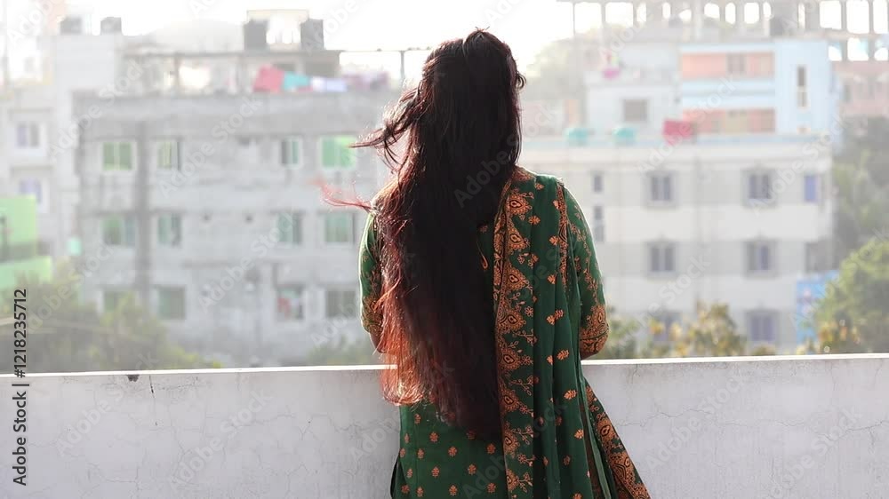 A Bengali woman, wearing a green block-printed salwar kameez, stands alone on a rooftop. Her long, reddish-black hair is gently moved by the breeze as she gazes out, lost in thought. Back view.