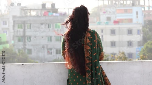 A Bengali woman, wearing a green block-printed salwar kameez, stands alone on a rooftop. Her long, reddish-black hair is gently moved by the breeze as she gazes out, lost in thought. Back view.