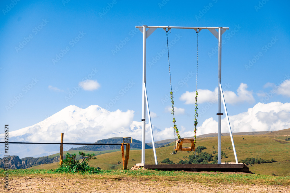 A swing is suspended from a metal pole in a field. The sky is clear and blue, and there are mountains in the background. The scene is peaceful and serene A swing is suspended from a metal pole in a field. The sky is clear and blue, and there are mountains in the background. The scene is peaceful and serene
