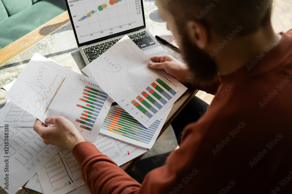 Naklejka premium Male accountant using laptop and paper documents to analyze financial investments on a data graph.