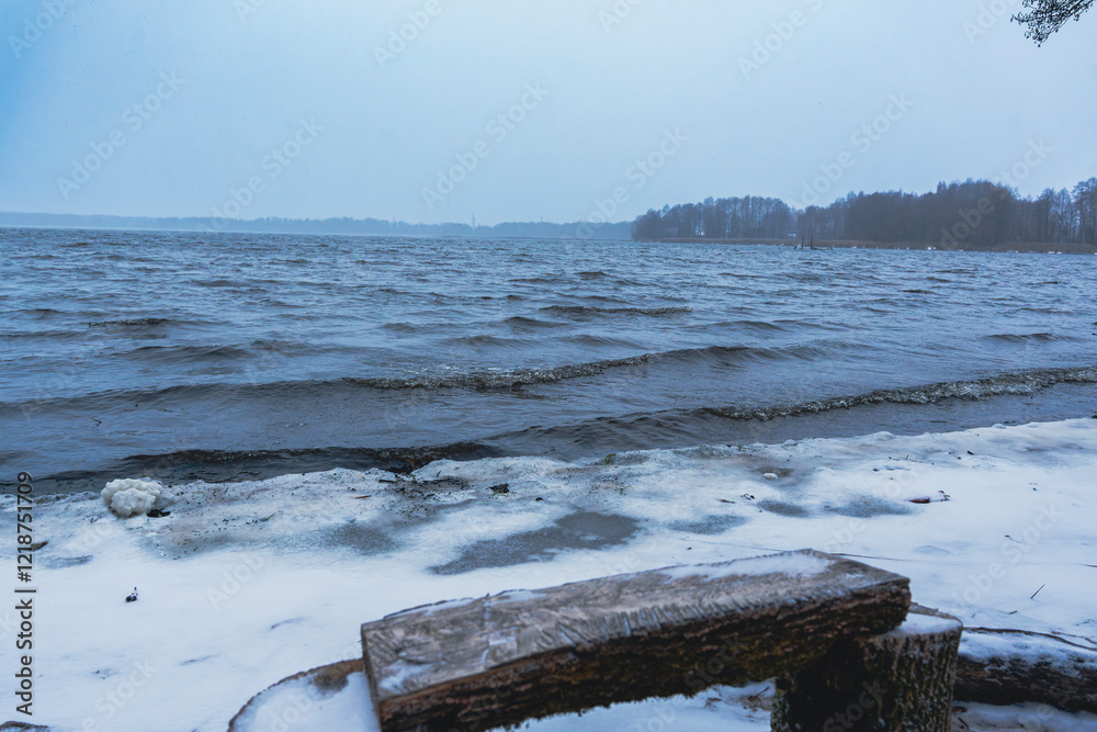 Fototapeta premium Snow-covered lakeshore with wooden bench and table on a winter day