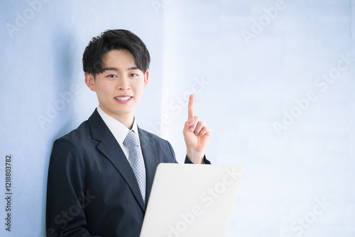 パソコンを持つ、指差しのポーズ若い20代男性　新入社員などのイメージ　笑顔のカメラ目線の水色背景