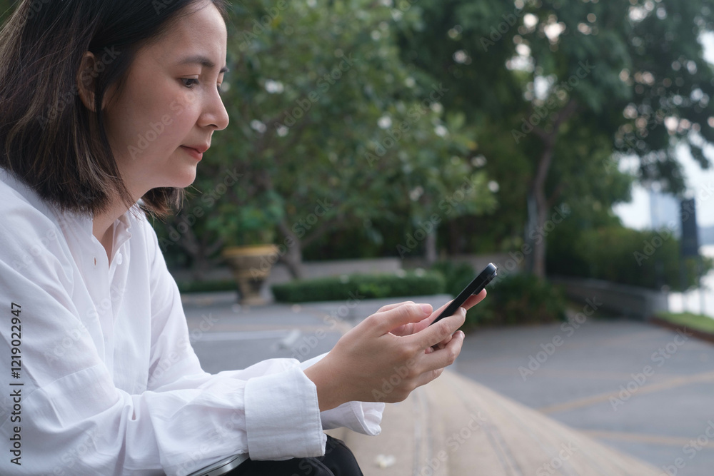 A woman sits outdoors in a park, engaged with her smartphone. Trees and pathways surround her, capturing a serene and modern lifestyle moment.
