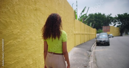 Teenager of African descent walking away along a bright yellow wall, back view, showcasing natural curly hair and casual style in an urban outdoor environment
