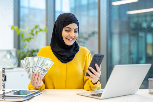 Joyful and successful woman in hijab sitting at desk inside office. Arab businesswoman holding money cash dollars and phone in hands, received online loan using banking app. photo