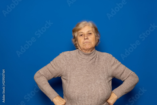 Caucasian woman in her 70s wearing a dark beige sweater with her arms akimbo and a serious and defiant expression. Isolated on blue studio background.