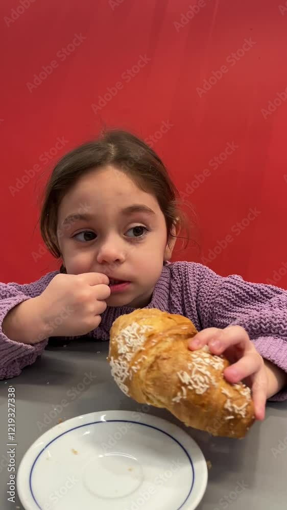 vertical video of a cute toddler girl with two pigtails in purple pullover eating fresh delicious croissant in caffe with red sofa vertical video of a cute toddler girl with two pigtails in purple pullover eating fresh delicious croissant in caffe with red sofa