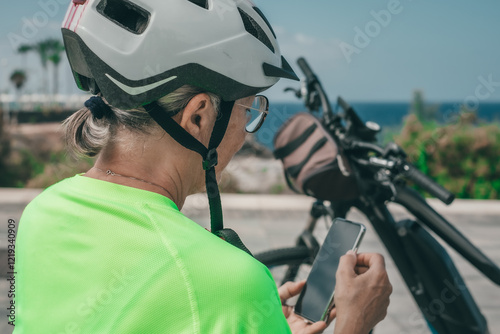 Back view of senior athletic woman with bicycle stops on a bench to answer the phone, elderly lady enjoying freedom, authentic healthy retirement lifestyle #1219340909