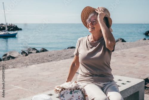 Happy carefree senior woman holding her hat sitting on a bench at seaside enjoying free time vacation and retirement. Old people and healthy lifestyle #1219341548