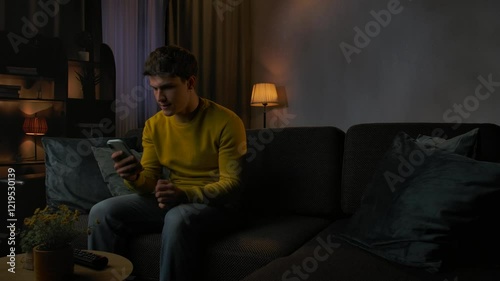 Young man in the evening after work relaxing at home, sits on the sofa in living room watching tv, holds smartphone anticipating getting good news, winning expression.