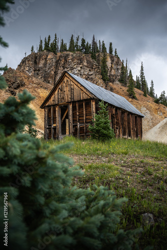 Abandoned wooden mine building nestled in a scenic mountain setting with dramatic rocky cliff and evergreen trees, evoking rustic charm and natural beauty.