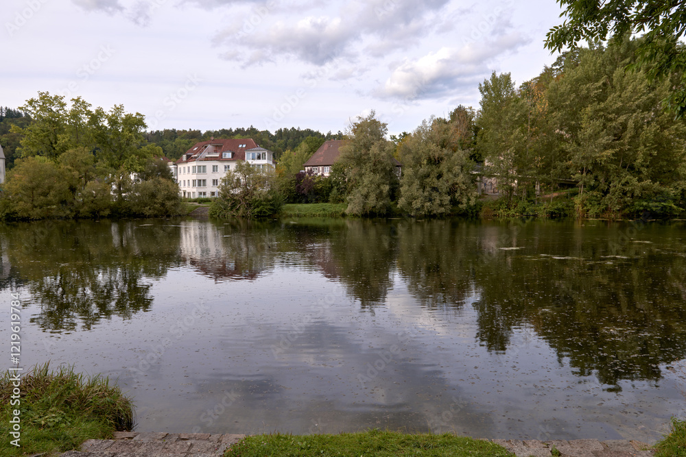 Obraz premium Serene River Landscape with Cozy Cottages Nestled in Lush European Greenery Under Dramatic Twilight Sky. Tranquil European Riverside Village at Dusk: Picturesque Houses Nestled Among Lush Greenery