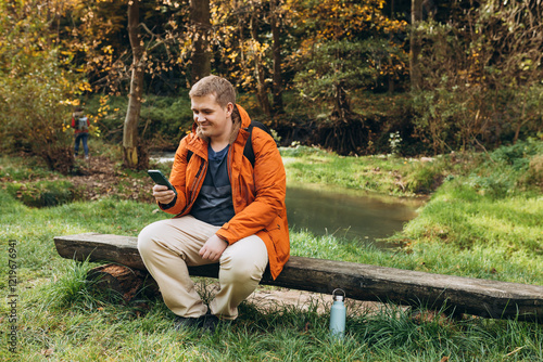 Obraz na płótnie Stylish man with backpack holding smart phone and relaxing in nature. Full body photo. Hiking active male lifestyle siting on tree trunk outdoors. High quality photo. Adventures. w salonie