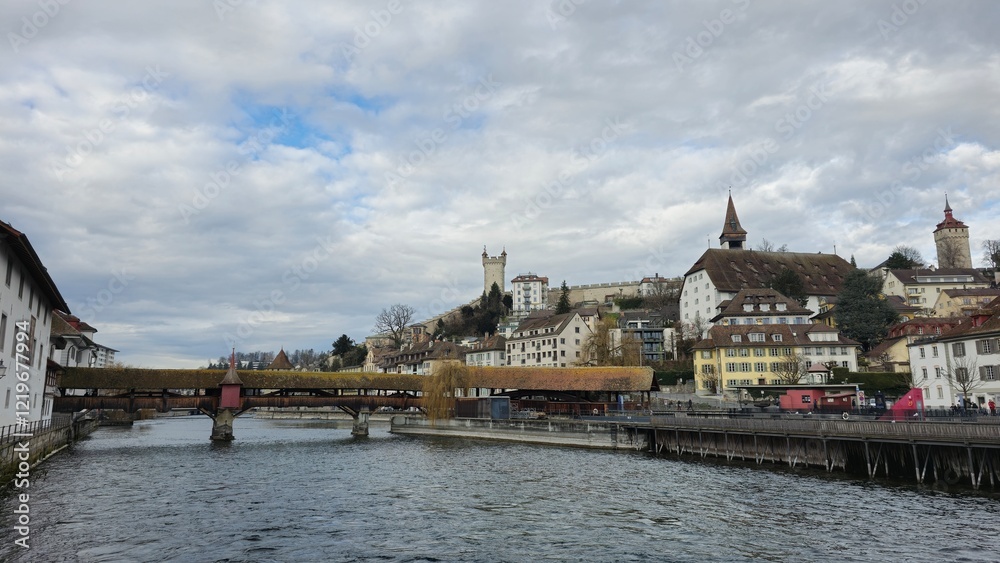 Obraz premium A scenic view of Lucerne with a wooden covered bridge spanning the water, historic buildings, towers, and a hilltop castle under a cloudy sky, creating a picturesque European landscape.