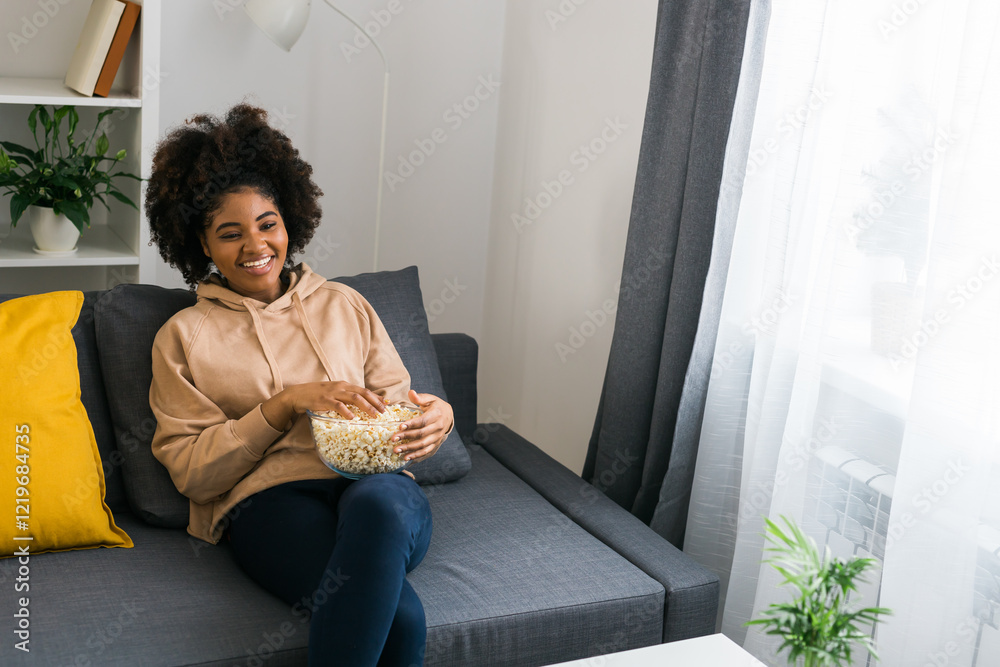 Young African American woman holds a bowl of popcorn while enjoying a television show. Bright smile radiates happiness and relaxation in a cozy home. TV, series and streaming service concept. Copy Young African American woman holds a bowl of popcorn while enjoying a television show. Bright smile radiates happiness and relaxation in a cozy home. TV, series and streaming service concept. Copy