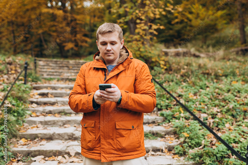 Adult 30s man hiker stand outdoors in a natural setting, smile while using his mobile phone outdoors. Hiking, lifestyle, adventure and tourism concept.