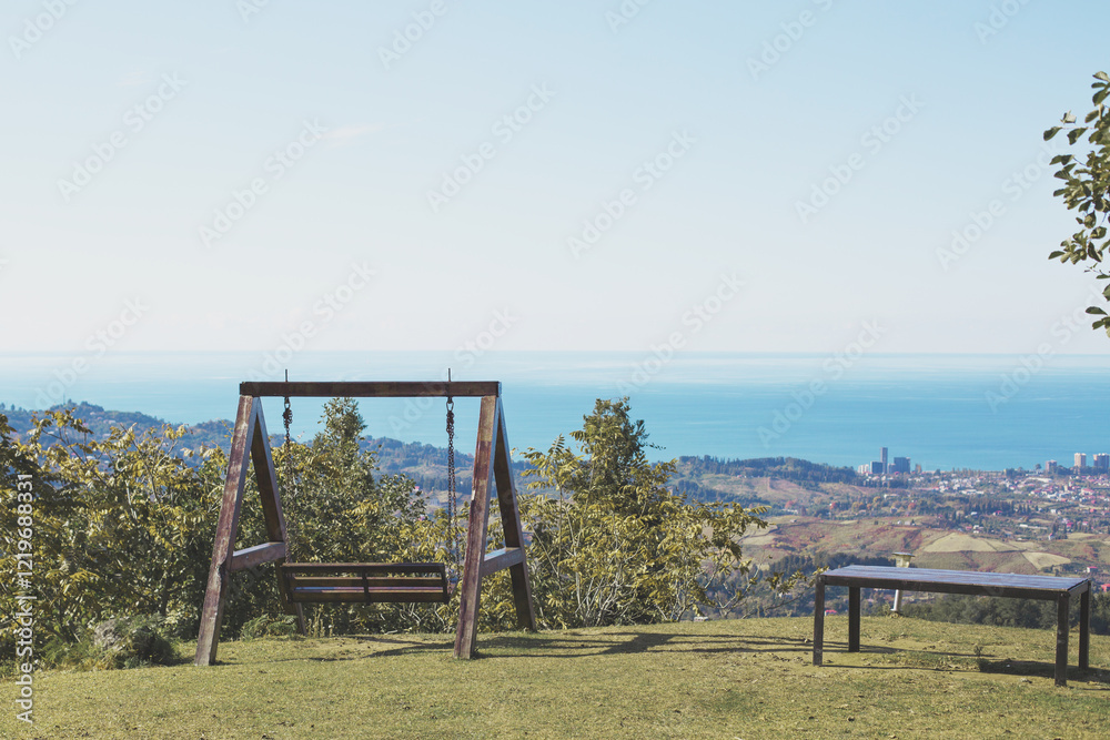 Wooden swing and bench on a grassy hilltop overlooking the sea and a distant city under a clear blue sky, evoking peace and simplicity Wooden swing and bench on a grassy hilltop overlooking the sea and a distant city under a clear blue sky, evoking peace and simplicity