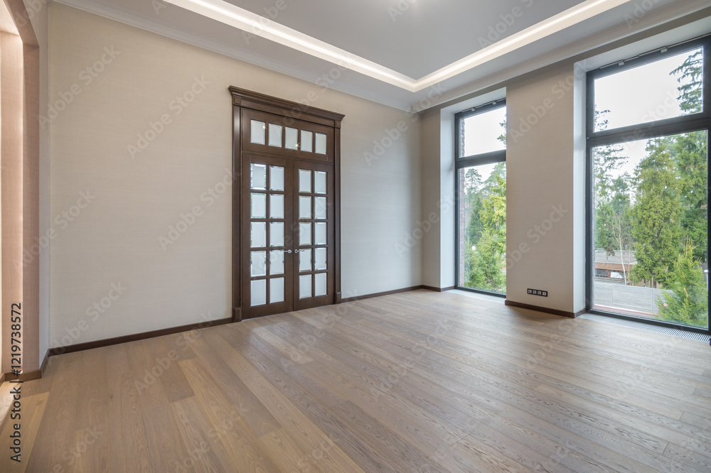 Obraz premium A spacious empty room with the final finish in a luxurious mansion. Brown door with frosted glass windows. Panoramic windows overlooking the green landscape. Unfurnished.