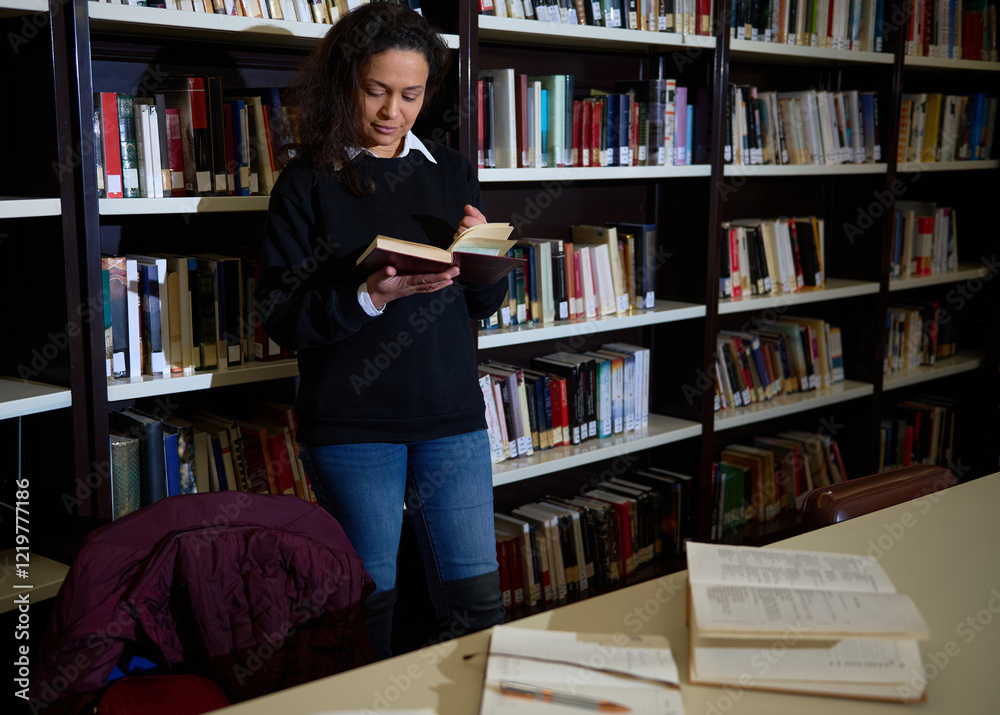 A woman stands in a library, engrossed in a book, surrounded by shelves of literature, and studying at a desk. A woman stands in a library, engrossed in a book, surrounded by shelves of literature, and studying at a desk.