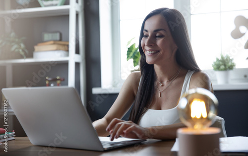 A smiling young woman working on her laptop at a modern home office. She’s focused and enjoying her work with a relaxed vibe, surrounded by plants, books, and a stylish lightbulb lamp. Free Download A smiling young woman working on her laptop at a modern home office. She’s focused and enjoying her work with a relaxed vibe, surrounded by plants, books, and a stylish lightbulb lamp. - GFXLoad