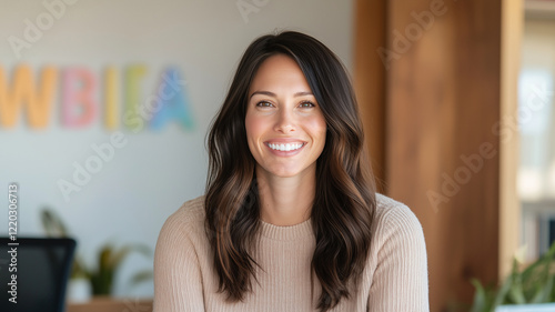 smiling woman with long, wavy hair, wearing beige sweater, sitting in modern office environment. background features colorful letters, creating cheerful atmosphere