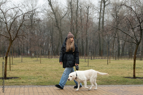 A young woman walks her yellow Labrador retriever in a quiet park, surrounded by budding trees and soft grass. The day is overcast, hinting at early spring
