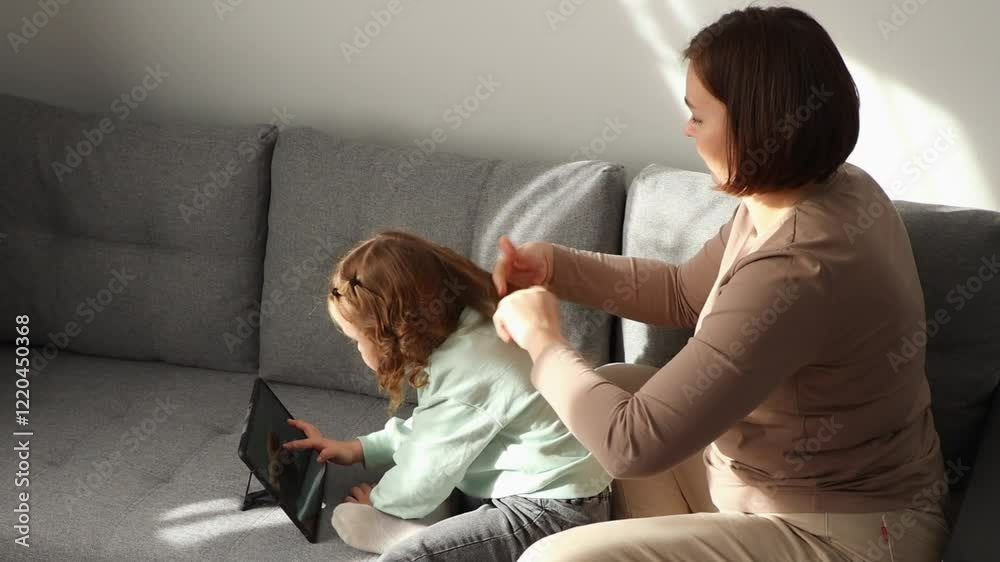 Side view of Caucasian little girl is watching cartoons on her tablet while adult woman braids her hair. Mother and daughter spend time together Side view of Caucasian little girl is watching cartoons on her tablet while adult woman braids her hair. Mother and daughter spend time together