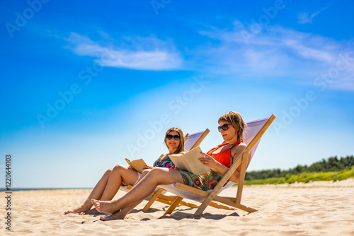 Summer holidays on beach with book. Two mature women reading books sitting on deckchairs on white sandy beach in summertime #1220644103