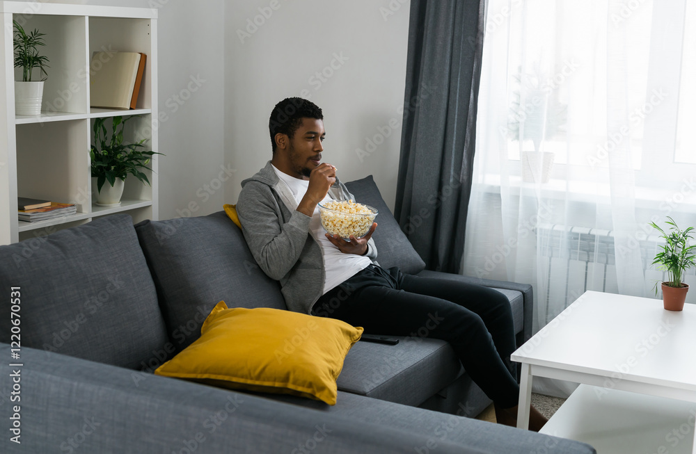 African American man sits alone on the couch and enjoying popcorn while watching television and streaming service African American man sits alone on the couch and enjoying popcorn while watching television and streaming service