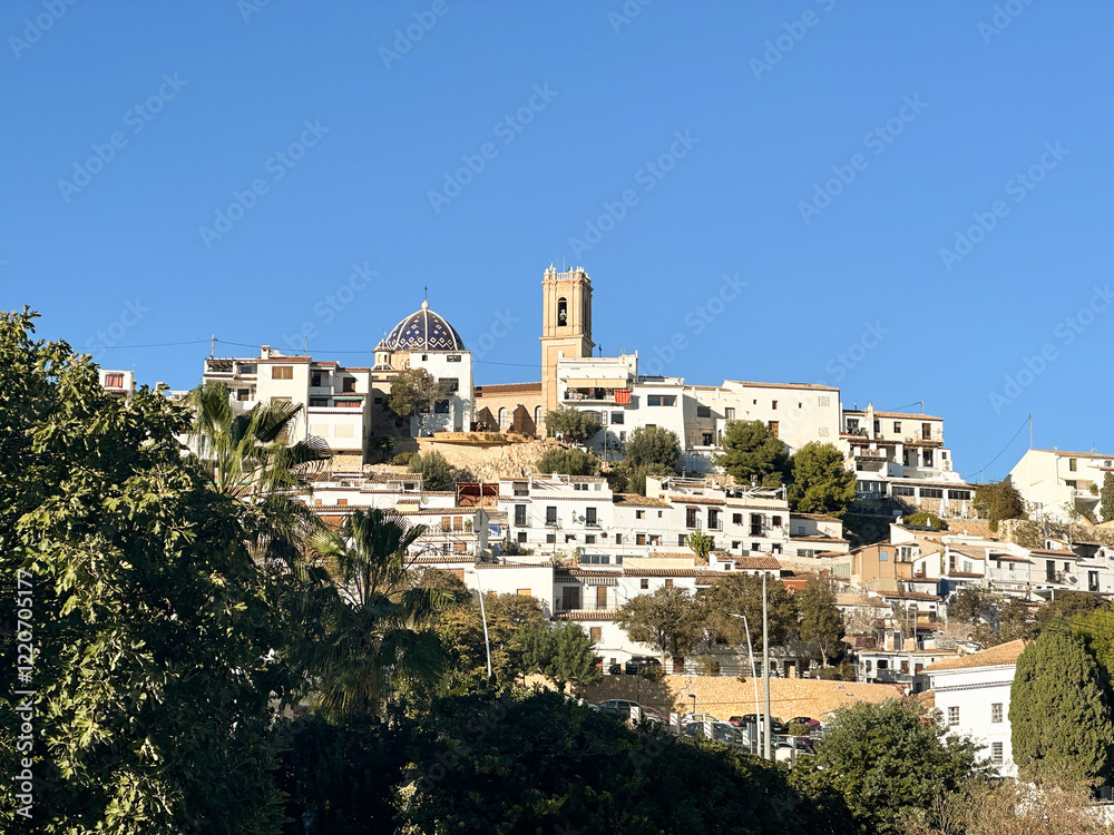 Obraz premium Scenic view of Altea village with whitewashed houses, a blue-tiled dome, and a bell tower on a hillside under a clear blue sky