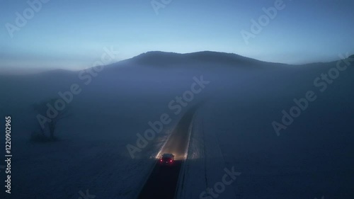 Eerie winter landscape with car on lonely road and mountains skyline aerial view. Automobile driving through morning fog on empty rural highway drone tracking shot