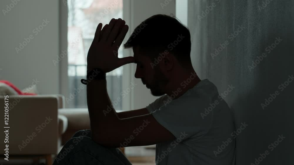 Depressed young man sitting on floor in dark room. Silhouette of young male suffering from loneliness and anxiety, covering his face with hands. Person with mental disorder