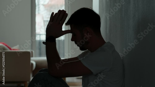 Depressed young man sitting on floor in dark room. Silhouette of young male suffering from loneliness and anxiety, covering his face with hands. Person with mental disorder