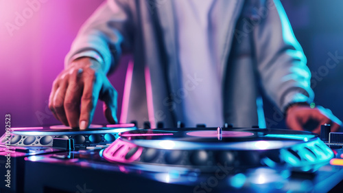 DJ spinning vinyl records with vibrant lights pulsing in background creates energetic atmosphere. focus is on DJ hand adjusting controls, enhancing lively scene