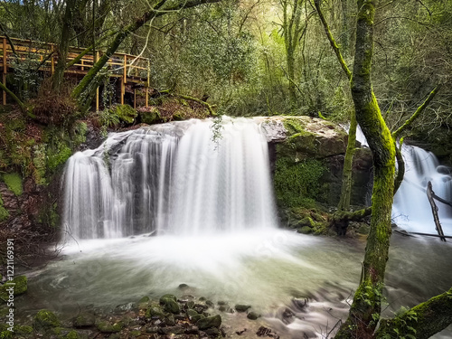 Waterfall in a lush forest with a wooden observation deck above, surrounded by moss-covered rocks and trees, creating a serene and picturesque scene in Pindelo, Outeiro, Oliveira de Azemeis, Portugal photo