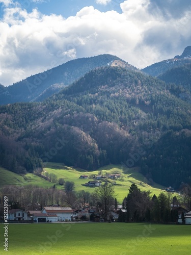 A picturesque view of Aschau im Chiemgau in Bavaria, showcasing rolling green meadows, a quaint village, and forested mountains under a dramatic cloudy sky. photo