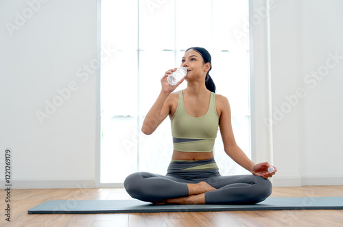 Pretty Asian woman with exercise clothes drink water from bottle and sit on yoga mat and stay in front of day light through glass window.
