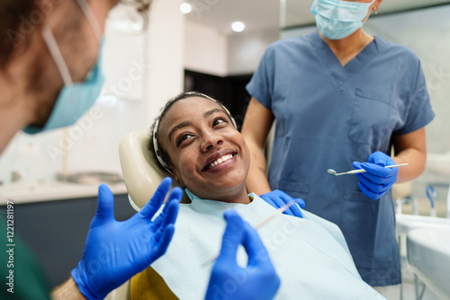 A Black woman smiles warmly while sitting in a dental chair. She is attended by a diverse dental team, including a male and a female, both wearing blue scrubs and gloves. The setting is modern.
