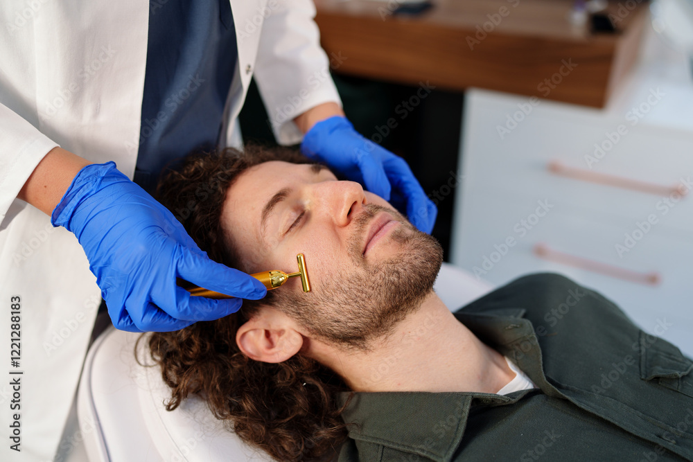 Fototapeta premium A Caucasian male enjoys a rejuvenating facial treatment using a gold beauty bar. A professional beautician in gloves performs the relaxing skincare routine in a modern spa setting.