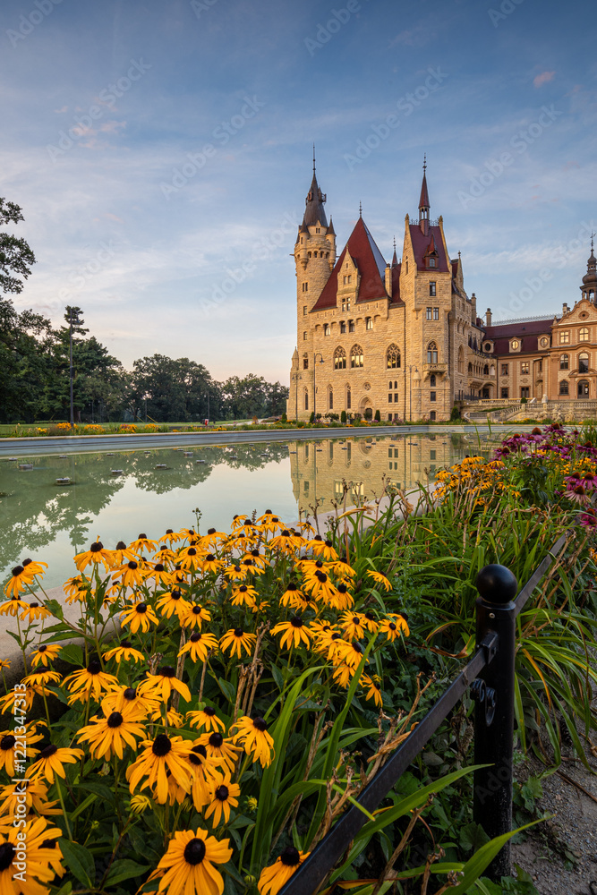 Majestic Moszna Castle on a warm summer morning, surrounded by lush greenery and glowing in soft golden sunlight, reflecting a fairytale-like ambiance perfect for travel and architecture themes.