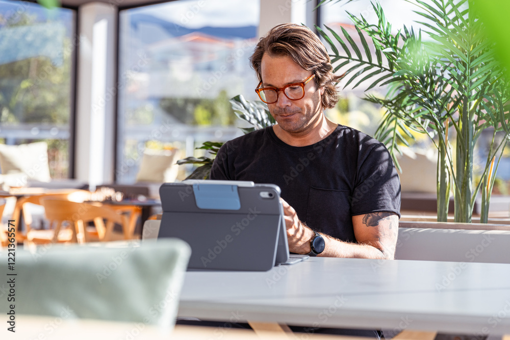 A man is sitting at a table working with his tablet and looking serious and concentrated A man is sitting at a table working with his tablet and looking serious and concentrated
