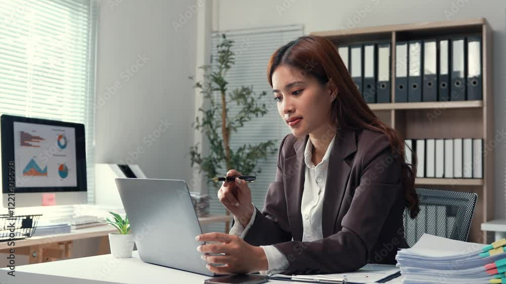 Young businesswoman working intently at office desk, using laptop and smartphone while holding pen, analyzing screen with focused professional demeanor Young businesswoman working intently at office desk, using laptop and smartphone while holding pen, analyzing screen with focused professional demeanor