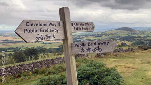 A wooden directional signpost for The Cleveland Way and public bridleway stands in a panoramic rural setting with open fields and hills in the background - Osmotherley, North Yorkshire, UK photo