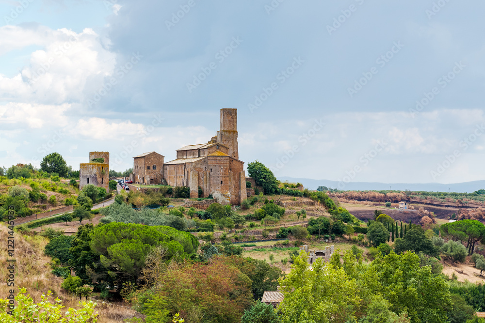 Fototapeta premium Church of San Pietro and two Etruscan towers rising on a hillside in Tuscania, Italy, with lush vegetation, trees, and a cloudy sky creating a picturesque landscape