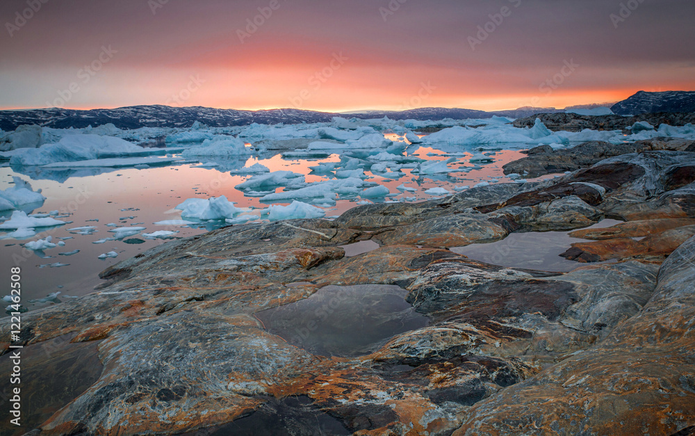 Obraz premium Rocky coastline and floating ice at sunset. East Greenland, Tasiilaq. Rocky shoreline, the water is covered in ice, and the sky is a beautiful orange and pink color.