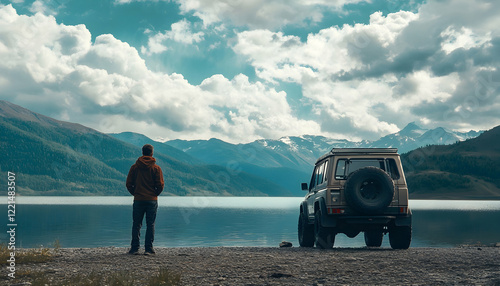 Middle-aged man standing near off-road car on the lake shore, backside view, looking at mountains in the distance. Cloudy sky. Off-road travel, journey, overlanding concept