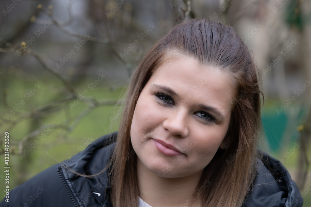 A young woman in a black jacket standing next to a tree, smiling softly, with a natural outdoor setting and bare branches in the background. A young woman in a black jacket standing next to a tree, smiling softly, with a natural outdoor setting and bare branches in the background.
