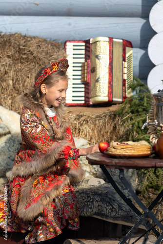 A blonde girl in a national Russian costume on the Maslenitsa holiday. A beautiful russian girl in a national costume made of a fur cape and kokoshnik on the background of a hayloft. 