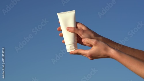 Female hands squeeze cream or lotion from a white mockup tube onto the palm of her hand against a blue sky background. Concept of beauty care products, sun protection and skin hydration.