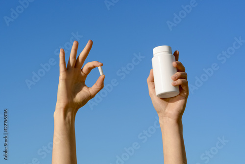 Female hand holding white capsule pill and white mockup jar of pills on blue sky background. Concept of pharmacy, medicine and natural dietary supplements for health support and treatment